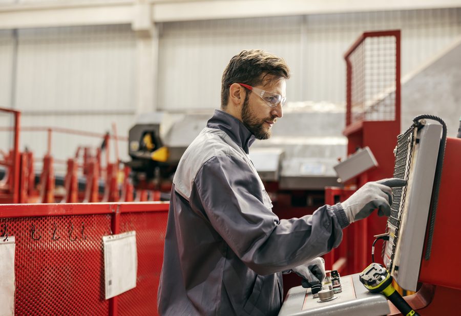 Mann in Arbeitskleidung mit Schutzbrille bedient eine Maschine in einer Fertigungshalle