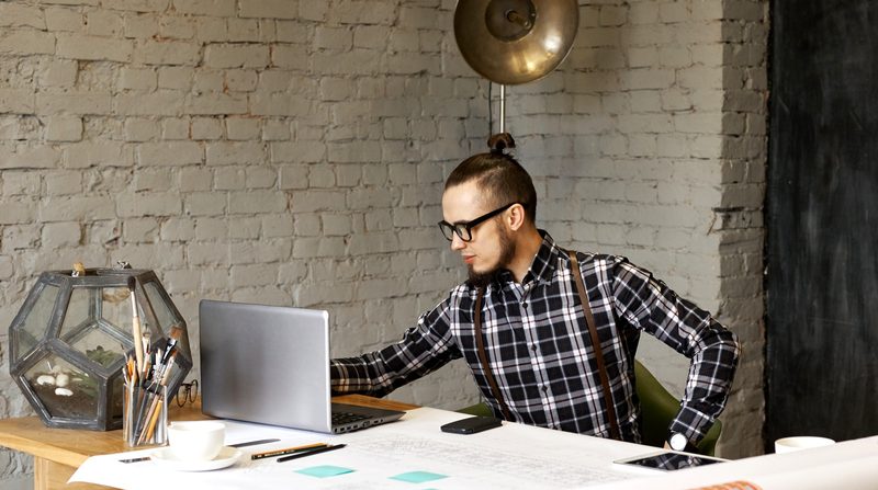 Mann mit Hornbrille und Hosenträgern arbeitet am Laptop in einem Loft mit Backsteinwand